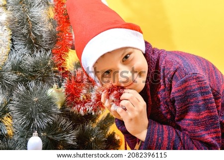 A boy wearing a red Santa Claus hat decorates a Christmas tree with festive decorations and toys.