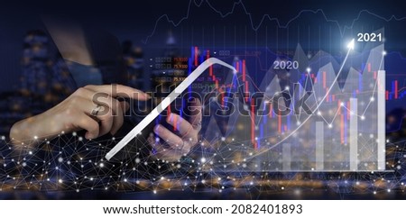 Financial charts showing growing revenue In 2021. Hand touch white tablet with digital hologram growth graph chart sign on city dark blurred background. Business growth concept year 2021