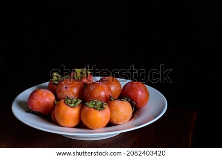 A plate of ripe persimmons stands on a wooden table against a dark background