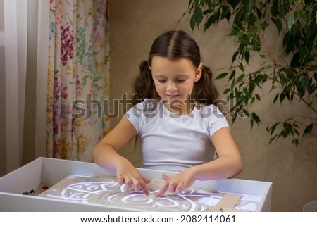Pretty child girl paint an illustration by finger with sand on light table at home. Sand therapy concept.