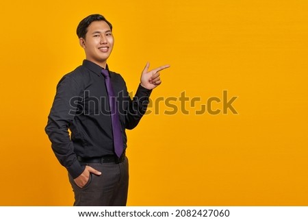 Smiling young handsome businessman pointing finger away at copy space on yellow background