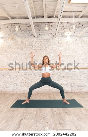beautiful woman doing goddess yoga pose, wearing sportswear, green pants, white top, indoor full length, yoga studio. High quality photo