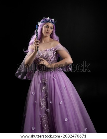 Full length portrait of girl wearing purple fantasy dress with fairy wings and crown. pose with gestural hand movements as if casting magical spells.  isolated on dark studio background 