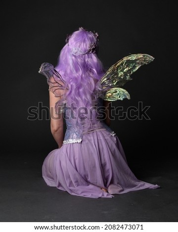 Full length portrait of girl wearing purple fantasy dress with fairy wings and crown. Sitting pose with gestural hand movements as if casting magical spells.  isolated on dark studio background 