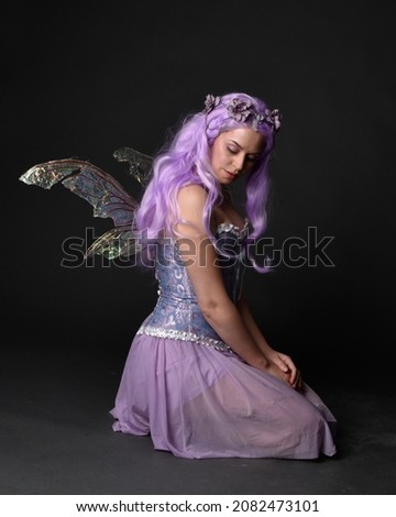 Full length portrait of girl wearing purple fantasy dress with fairy wings and crown. Sitting pose with gestural hand movements as if casting magical spells.  isolated on dark studio background 