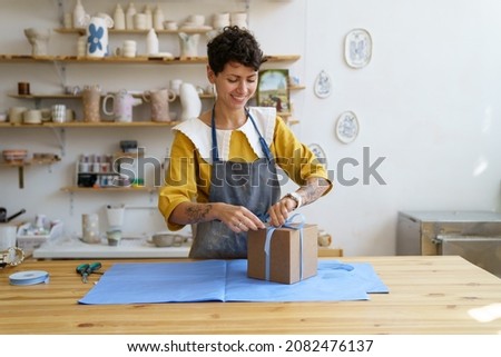 Woman seller in craft crockery and handmade pottery shop packing gift box for customer. Self-employed ceramist prepare parcel with handicraft ceramics for delivery. Artist decorate present in studio Royalty-Free Stock Photo #2082476137