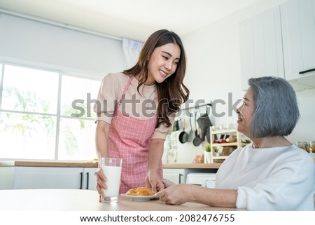 Asian lovely family, young daughter prepare breakfast for older mother. Attractive female wear apron bake croissant serve with milk to senior elderly mom sitting on eating table in kitchen at house. Royalty-Free Stock Photo #2082476755