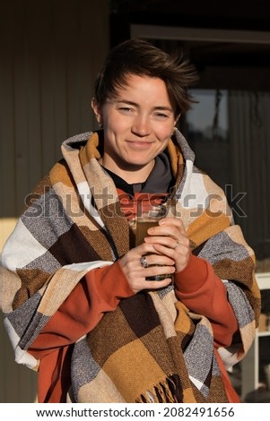 Beautiful girl resting and drinking juice sitting in autumn garden in chair wrapped in a woolen plaid blanket. copy space
