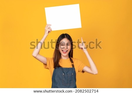 Happy young Asian woman wears an apron and holding a blank paper with smiling face and looking at the orange background. for advertising signs.