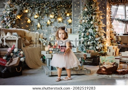 A little girl in an elegant dress plays with a colorful train toy in a room decorated for Christmas