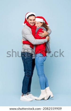 Full length portrait of happy Caucasian couple in Christmas outfits huging each other in light blue isolated studio background