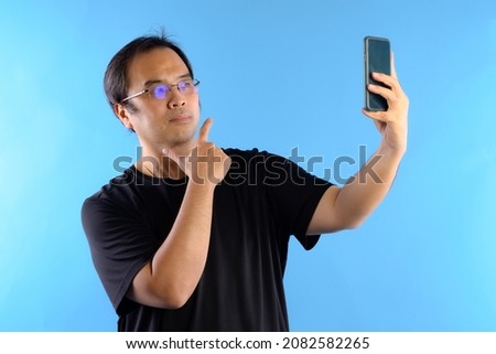 Asian man wore glasses and wear black t-shirts. Using mobile To take a picture  or selfie of himself isolated on blue background. 