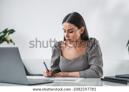 Businesswoman wearing formal dress is making notes or sign a contract. Office workplace with desk and notebook in the background. Concept of working process of director