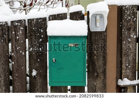 Snowy green mailbox and a ringing button at a wooden fence.