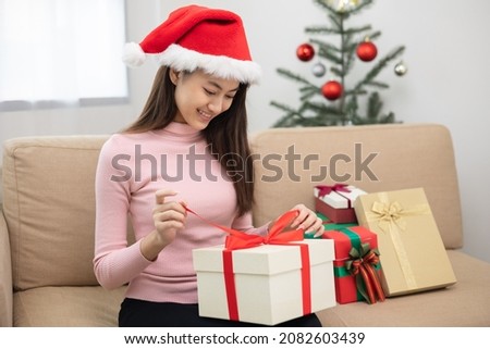 Beautiful Asian woman unpack the present gift box pull ribbon guess what inside the box. Young Happy cute girl celebrate in christmas holiday festival with many gift box sitting on sofa at home