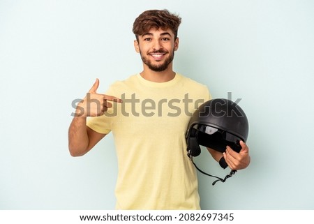 Young mixed race man holding motorcycle helmet isolated on blue background person pointing by hand to a shirt copy space, proud and confident Royalty-Free Stock Photo #2082697345