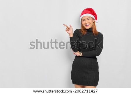 Smiling young Asian woman in Santa Claus hat pointing finger at copy space isolated over white background