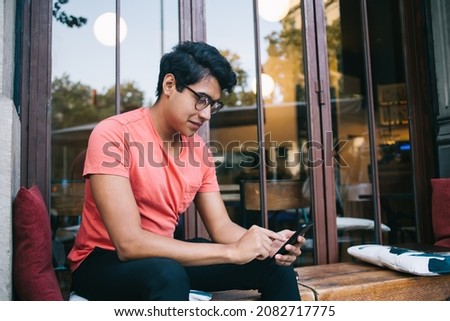 Side view of young male blogger connecting to 4g internet wireless for making web booking via cellphone application, handsome Latin user installing mobile app during social networking on leisure