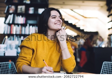 Smiling female employee in warm outfit looking away and thinking about startup assignment while sitting in cozy library during work