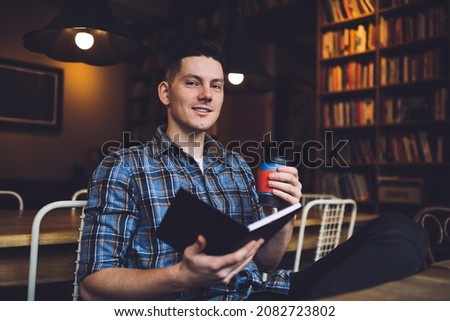Portrait of male student with disposable takeaway cup holding informative literature book for analyzing education knowledge during exam preparation in public library, intelligent schooler studying