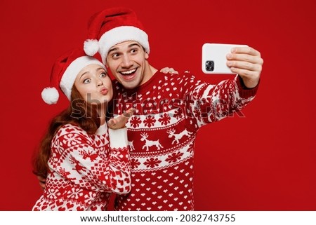 Young couple friends two man woman in sweater hat doing selfie shot on mobile cell phone blow air kiss isolated on plain red background. Happy New Year 2022 celebration merry ho x-mas holiday concept
