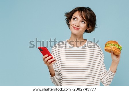 Young minded happy woman in striped shirt using mobile cell phone hold burger count calories browsing internet think isolated on plain pastel light blue background studio People lifestyle food concept