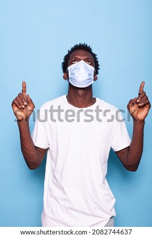 Casual man with face mask against coronavirus pointing up with index fingers. Young adult wearing protection for covid 19 virus pandemic while doing sign with hands in studio.