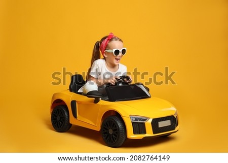 Cute little girl driving children's electric toy car on yellow background Royalty-Free Stock Photo #2082764149