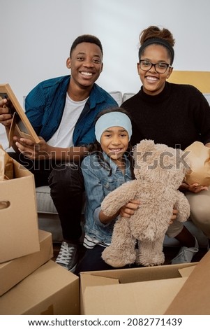 Loving family sits on couch in living room unpacks multitude of boxes standing around them after moving to new apartment they pull out old memorabilia parents with their daughter smile at camera