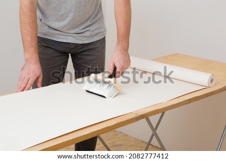 Young adult man hand using brush and applying glue on wallpaper sheet on wooden table. Closeup. Repair work of home.  Royalty-Free Stock Photo #2082777412