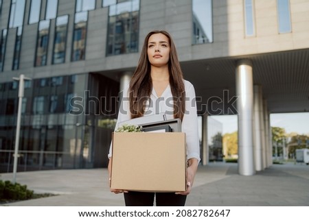 Woman stands in front of glass modern building of corporation office where she worked dismissal from position girl with sad uncertain face looks ahead holding box with packed things unemployment Royalty-Free Stock Photo #2082782647