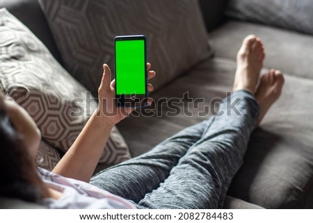 A young woman sitting on the couch is using a smartphone with a pre-configured green screen.