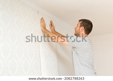 Young adult man hands applying new wallpaper on white wall. Closeup. Repair work of home. Side view. Royalty-Free Stock Photo #2082788263