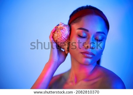 young woman with closed eyes pressed seashell to her ear enjoy and dreams with copy space in neon light. Summer vacation, relaxation concept