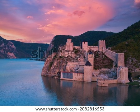 Aerial, across lake view of the medieval fortress Golubac over  Danube river. Fortress towers illuminated by pink light. Sunset, pink and red clouds sky. Outdoor and traveling theme. Serbia. Royalty-Free Stock Photo #2082816553