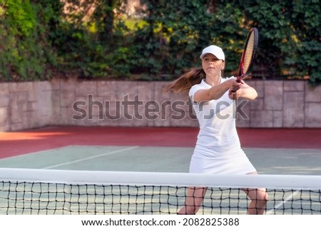 a woman in a white dress and a baseball cap plays lawn tennis on a tennis court in summer, hits the ball with a racket or makes a pitch, sports and recreation