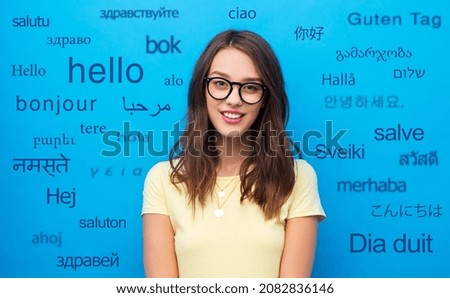 education, school and people concept - smiling young woman or teenage girl in yellow t-shirt and glasses over greeting words in different foreign languages on blue background