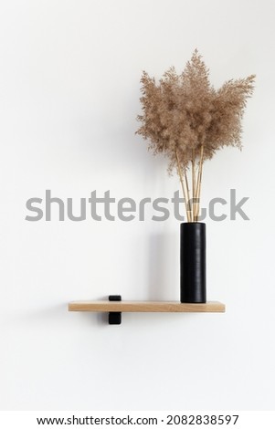 Vertical photo of short wooden shelf with black bracing and scandinavian style vase with dried plants over white wall background