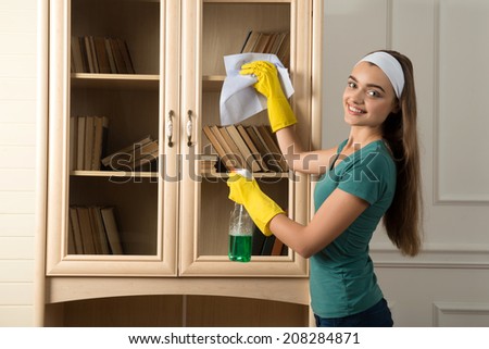Half-length portrait of nice smiling dark-haired housemaid standing aside in white fillet dusting the book shelf and looking at us
