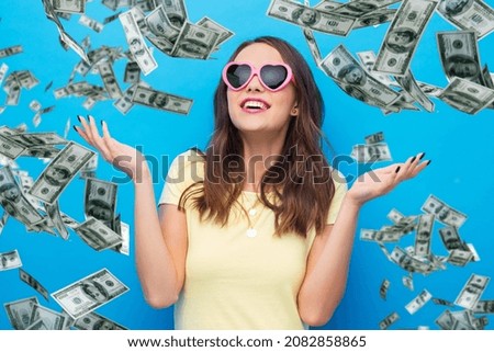 summer, valentine's day and people concept - smiling young woman or teenage girl in yellow t-shirt and heart-shaped sunglasses over dollar money rain on bright blue background Royalty-Free Stock Photo #2082858865