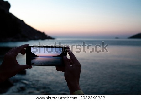 woman hands holding mobile phone at sunrise over sea bay. Taking photo of sunset over rocks in water by mobile phone. Selective focus on smartphone