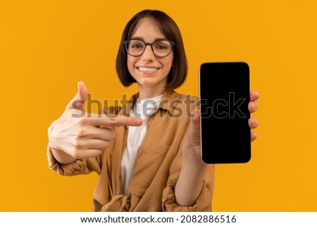 Great app. Excited woman pointing at smartphone blank screen, demonstrating free space for mobile app or website design, standing on yellow background, mockup