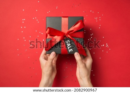 First person top view photo of hands holding black giftbox with red ribbon bow and tag shiny sequins on isolated red background with text on pricetag