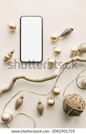 Modern smart phone with copy space screen for your content lying on surface surrounded with New Year decor, tiny cute gold baubles, tree branch and wrapping rope. Christmas arrangements