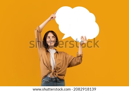 Happy young woman holding empty speech bubble over her head on yellow studio background, free space for design. Excited caucasian woman with blank word box, mockup