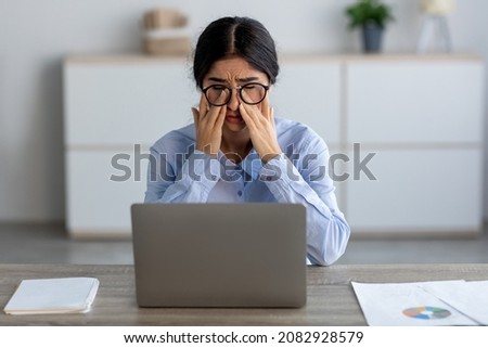 Tired sad young indian business woman suffering from headache, fatigue, rubs eyes at workplace with laptop in office or home interior. Dry eyes, stress, deadline, pain and ill. Disease and migraine Royalty-Free Stock Photo #2082928579