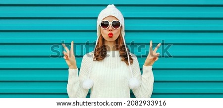 Winter portrait of beautiful young woman blowing her lips sending air kiss wearing a white knitted sweater, hat on blue background