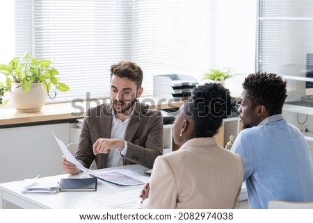 Young bearded lawyer examining contract or tax paper while giving consultation to young couple Royalty-Free Stock Photo #2082974038