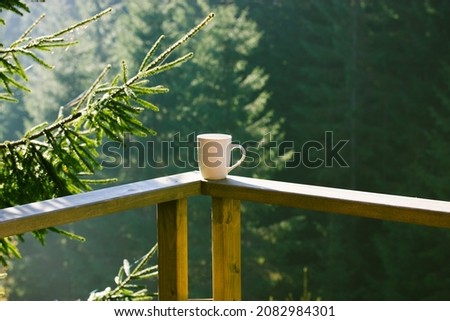 White ceramic cup of tea, coffee beverage stands on wooden balcony railing against green coniferous forest background, tall pines, firs in woods. Breakfast in the mountains. Spruce branch in sunlight. Royalty-Free Stock Photo #2082984301