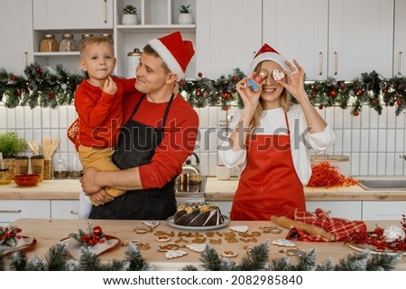 Merry Christmas and Happy Holidays. Family preparation holiday food in cosy kitchen at home. Mother in santa hat closes her eyes with gingerman. Father in santa hat holds cute little son in his arm